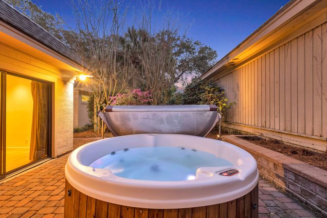 Cozy backyard hot tub on a brick patio at twilight, round jetted spa steaming beside a lit sliding glass door with garden plants and wooden fence.