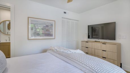 Bright modern bedroom interior with white bedding and patterned throw, wooden six-drawer dresser, wall-mounted flat-screen TV, framed artwork, louvered closet doors and an en-suite bathroom visible through the doorway.