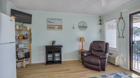 Coastal-themed beach cottage living room with a brown leather recliner by a sliding glass door and balcony water view, pale blue walls with nautical art and starfish accents, wall-mounted TV, wooden shelving and mini fridge, small black cabinet and light wood floors.
