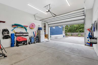 Spacious attached two-car garage with open door to driveway and blue-sided house exterior; white walls neatly organized with beach and pool gear—pool noodles, wagons, folding chairs, cooler and stroller—on a clean concrete floor.