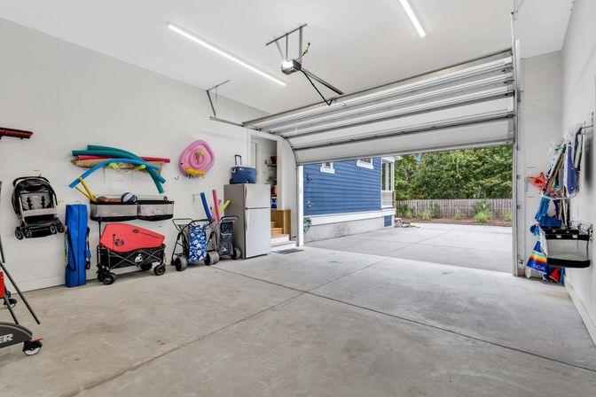 Spacious attached two-car garage with open door to driveway and blue-sided house exterior; white walls neatly organized with beach and pool gear—pool noodles, wagons, folding chairs, cooler and stroller—on a clean concrete floor.
