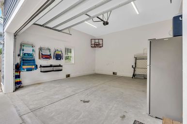 Tidy residential two-car garage with concrete floor and white walls, colorful wall-mounted beach chairs and hanging organizers, small window, wall-mounted mini basketball hoop, folded table and stainless refrigerator.