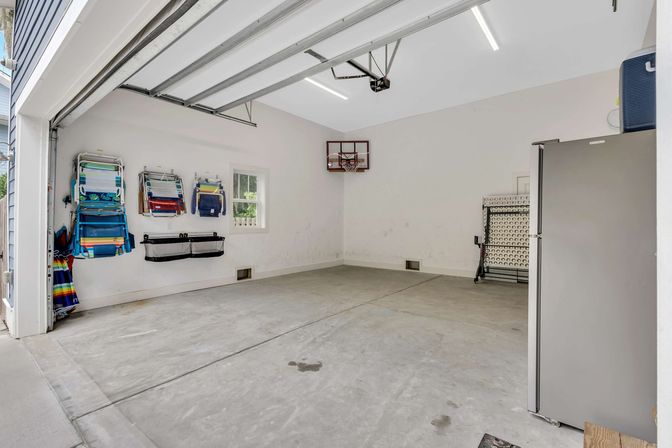 Tidy residential two-car garage with concrete floor and white walls, colorful wall-mounted beach chairs and hanging organizers, small window, wall-mounted mini basketball hoop, folded table and stainless refrigerator.