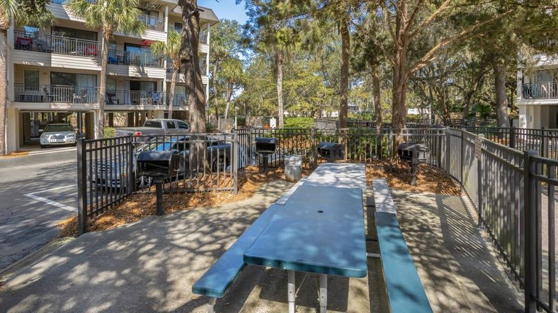 Sun-dappled outdoor picnic area with a blue picnic table and benches, several charcoal grills behind a metal fence in a leafy apartment courtyard with balconies and parked cars.