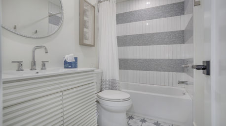 Bright modern residential bathroom with ribbed white vanity, chrome faucet and round mirror, toilet, patterned floor tiles, and bathtub/shower with gray-striped tile and white curtain.