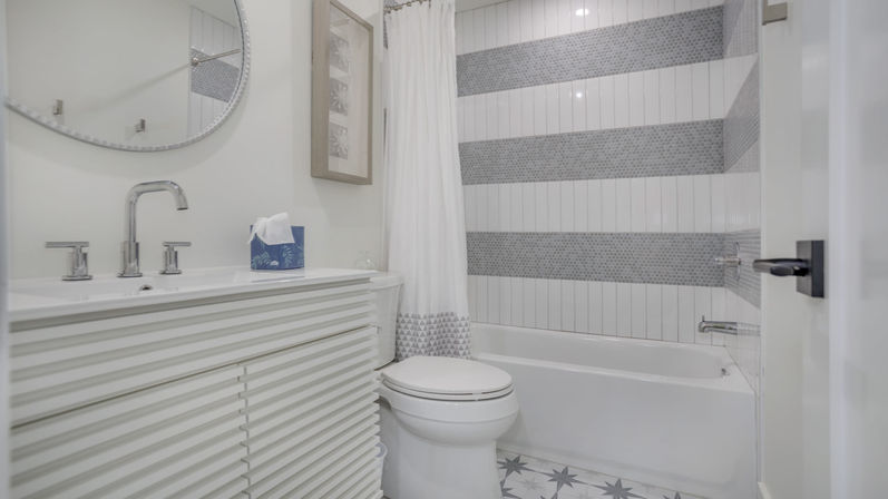 Bright modern residential bathroom with ribbed white vanity, chrome faucet and round mirror, toilet, patterned floor tiles, and bathtub/shower with gray-striped tile and white curtain.