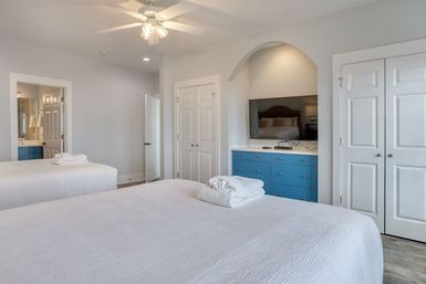 Bright modern guest bedroom with two white beds and folded towels, ceiling fan, TV in an arched alcove above a blue dresser, double closets and en suite bathroom doorway.