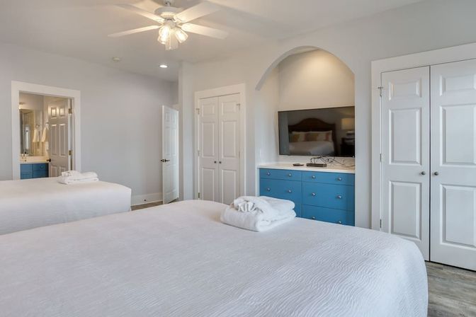 Bright modern guest bedroom with two white beds and folded towels, ceiling fan, TV in an arched alcove above a blue dresser, double closets and en suite bathroom doorway.