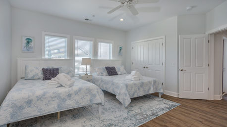 Bright coastal-style guest bedroom with two double beds in blue starfish quilts, folded robes on each bed, central nightstand and lamp, three shuttered windows, ceiling fan, and a patterned rug over wood floors.