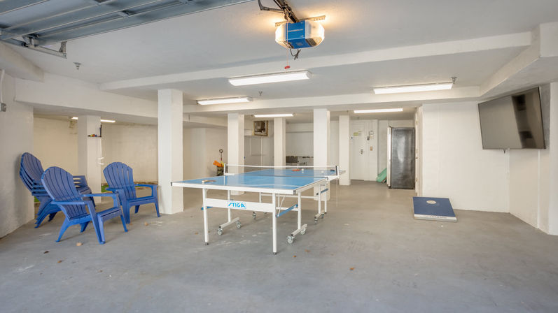 Brightly lit finished garage game room with a blue ping-pong table on casters, three blue plastic Adirondack chairs, a cornhole board, wall-mounted TV and concrete floor
