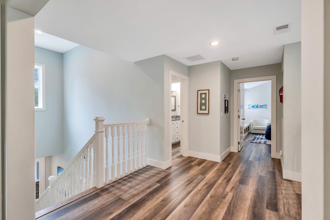 Bright second-floor landing in a contemporary home with warm wood-look flooring, white spindle railing, soft gray walls, recessed lighting, and doorways to a bathroom and bedroom.