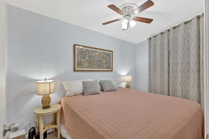 Cozy home bedroom interior with coral quilted bed, gray accent pillows, woven bedside lamps on round wooden tables, abstract framed art, patterned gray curtains and wooden ceiling fan.
