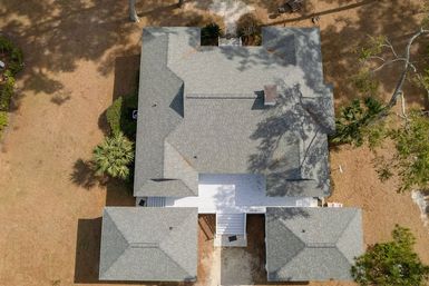 Aerial drone view of a gray-shingled gabled house with white rear deck, two detached outbuildings, dry yard and palm tree shadows