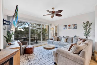 Bright coastal condo living room with a large gray L-shaped sectional, round wood coffee table, ceiling fan and wall-mounted TV, sliding glass doors to a balcony overlooking palm trees and neighboring resort-style building