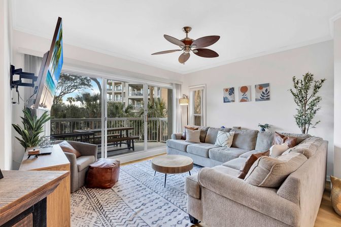 Bright coastal condo living room with a large gray L-shaped sectional, round wood coffee table, ceiling fan and wall-mounted TV, sliding glass doors to a balcony overlooking palm trees and neighboring resort-style building