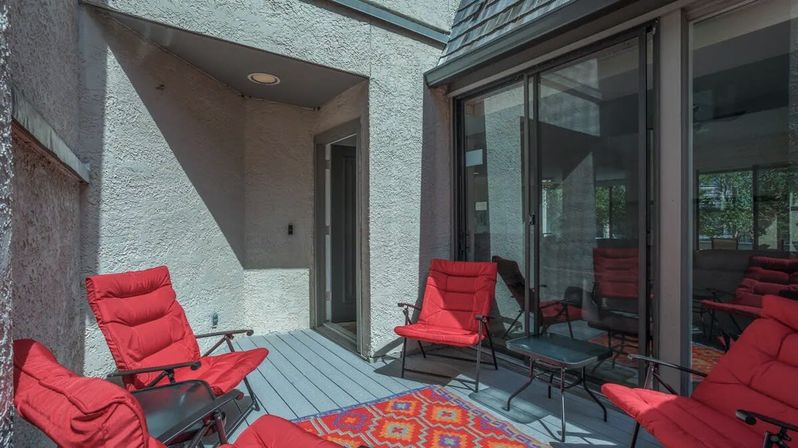 Covered patio with gray wood decking and sliding glass doors, furnished with four bright red cushioned lounge chairs, small black side tables, and a colorful geometric rug