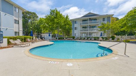 Resort-style outdoor swimming pool at a three-story apartment complex with curved deck, lounge chairs and umbrellas, balconies and trees under a sunny blue sky.