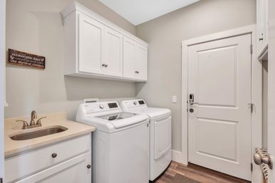 Modern residential laundry room with white top-load washer and matching dryer, white shaker cabinets, beige quartz countertop with sink and brushed-nickel faucet, neutral gray walls and wood-look flooring.