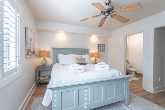 Bright coastal-style bedroom with light-blue wooden bed, white linens and stacked towels, matching nightstands and lamps, ceiling fan, plantation shutters and open door to a small ensuite bathroom.