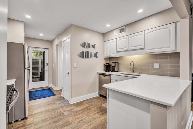 Bright coastal-inspired galley kitchen with white cabinetry and quartz breakfast bar, stainless steel appliances, beige subway-tile backsplash, wood-look flooring, striped fish wall art and a glass back door