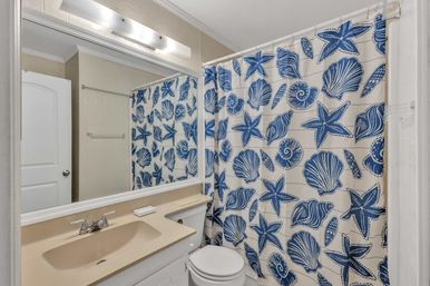 Beach-inspired small bathroom with beige sink and white vanity, large mirror and light fixture, white toilet, and blue seashell-and-starfish shower curtain