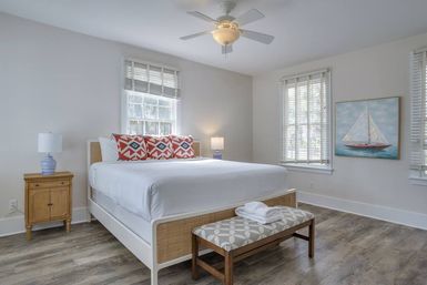 Bright coastal-inspired bedroom interior with white king bed, red patterned pillows, wooden nightstand and lamps, patterned bench at foot, sailboat wall art, large windows and ceiling fan