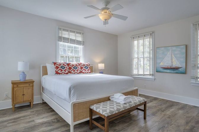 Bright coastal-inspired bedroom interior with white king bed, red patterned pillows, wooden nightstand and lamps, patterned bench at foot, sailboat wall art, large windows and ceiling fan