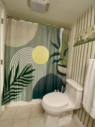 Small residential bathroom with white one-piece toilet, colorful abstract botanical shower curtain (green leaves, yellow sun and arched stripes), green striped wallpaper with lighthouse tile border, white towel and beige tile floor