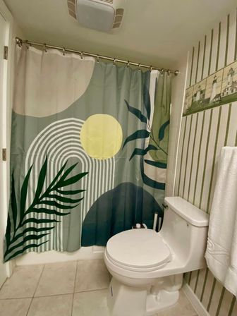 Small residential bathroom with white one-piece toilet, colorful abstract botanical shower curtain (green leaves, yellow sun and arched stripes), green striped wallpaper with lighthouse tile border, white towel and beige tile floor