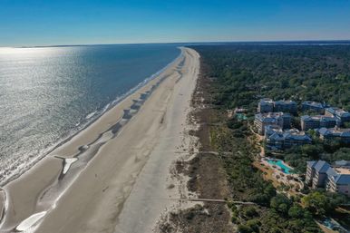 Aerial view of a long sandy beach with tidal sandbars and sparkling ocean beside a tree-lined coastline and seaside resort pools.