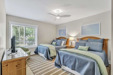 Bright guest bedroom with two twin beds in blue quilts and green throws, wooden headboards, nightstand with lamp, ceiling fan, striped rug, dresser with TV, and a large window overlooking trees