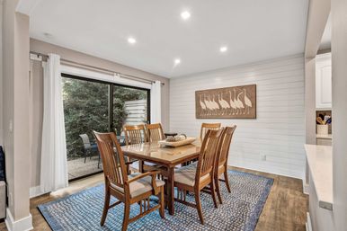 Cozy modern dining room with a wooden six-seat table and chairs on a blue patterned rug, white shiplap accent wall with framed bird artwork, recessed ceiling lights, and a sliding glass door opening to a leafy patio.