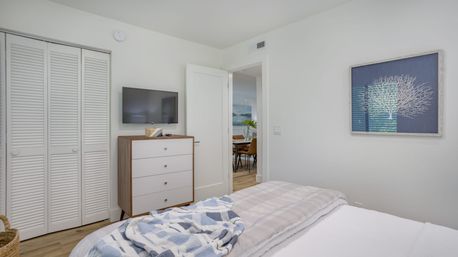Bright modern coastal-style bedroom interior with white walls, louvered closet doors, mid-century dresser under a wall TV, blue coral artwork, wood floors and layered plaid bedding with a folded blue throw, open door to dining area.