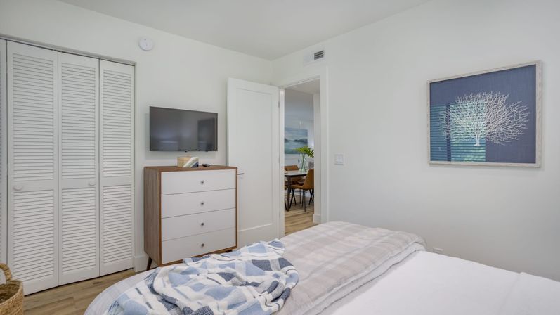 Bright modern coastal-style bedroom interior with white walls, louvered closet doors, mid-century dresser under a wall TV, blue coral artwork, wood floors and layered plaid bedding with a folded blue throw, open door to dining area.