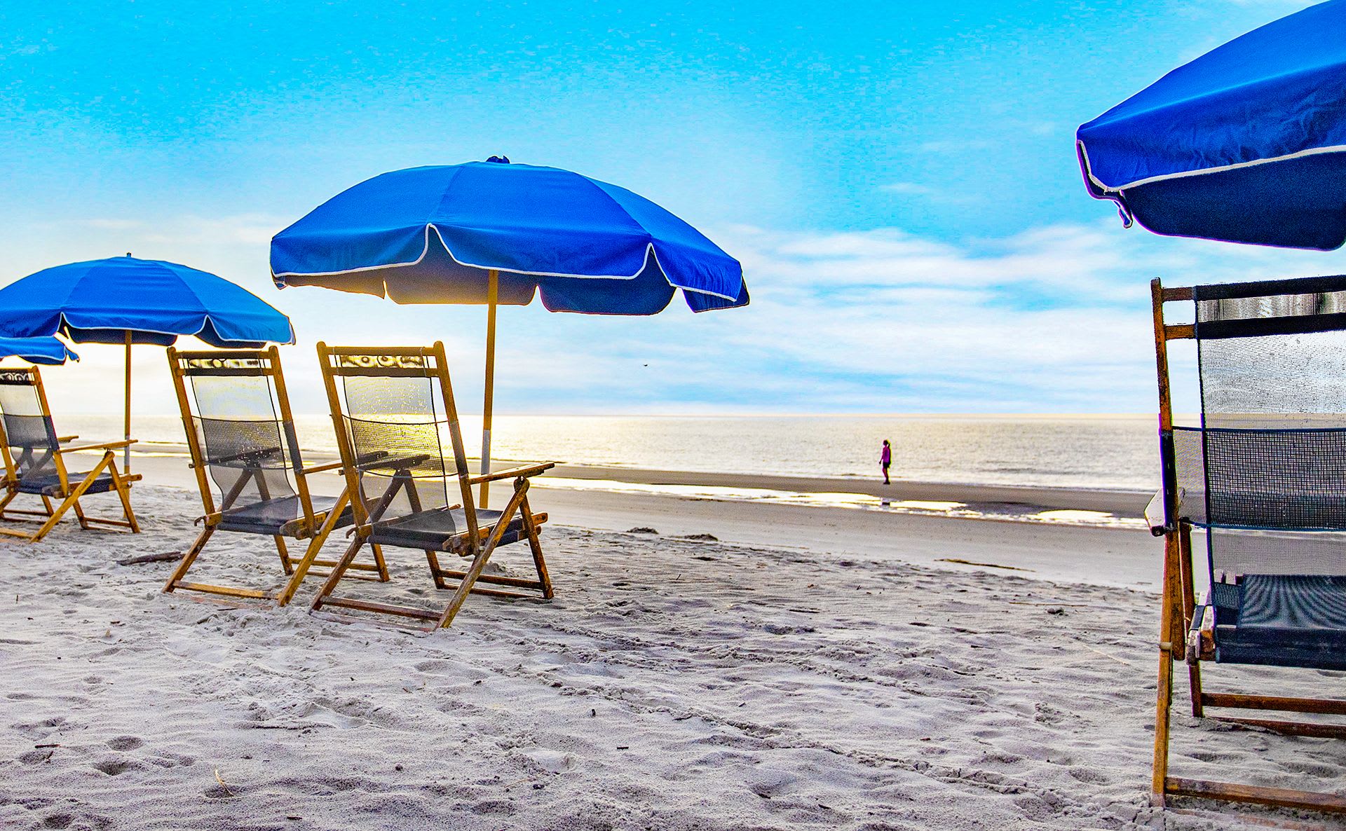 Sunlit sandy beach with wooden lounge chairs under bright blue umbrellas facing the calm ocean and a lone walker on the shoreline — a peaceful seaside relaxation scene.