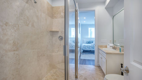 Bright modern ensuite bathroom with beige tiled walk-in shower and glass door, white vanity with mirror and sink, and a doorway opening to a sunny bedroom with patterned bedding.