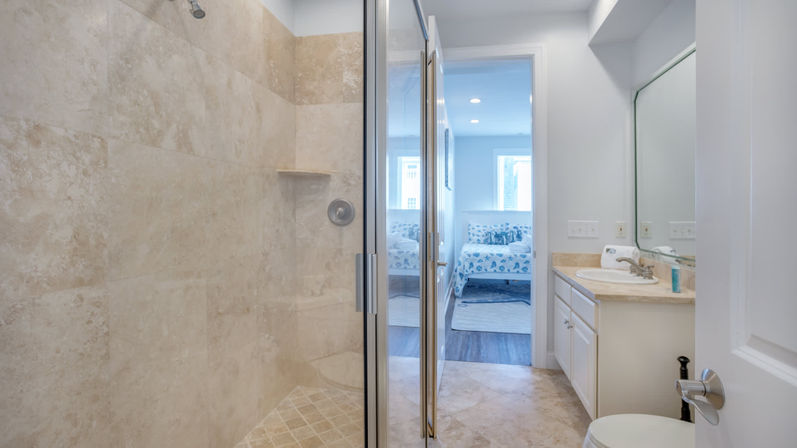 Bright modern ensuite bathroom with beige tiled walk-in shower and glass door, white vanity with mirror and sink, and a doorway opening to a sunny bedroom with patterned bedding.
