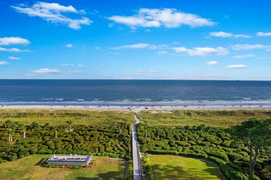 Aerial oceanfront view of a wooden boardwalk through green dunes to a sandy beach with small groups of beachgoers and a deep-blue sea under a bright, cloud-dotted sky