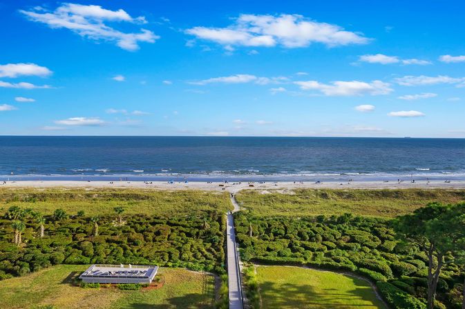 Aerial oceanfront view of a wooden boardwalk through green dunes to a sandy beach with small groups of beachgoers and a deep-blue sea under a bright, cloud-dotted sky
