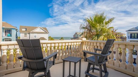 Ocean-view balcony with two dark Adirondack chairs and a small table behind a stone balustrade, overlooking palm trees, coastal homes, and the blue sea under a sunny sky.