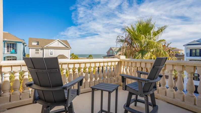 Ocean-view balcony with two dark Adirondack chairs and a small table behind a stone balustrade, overlooking palm trees, coastal homes, and the blue sea under a sunny sky.