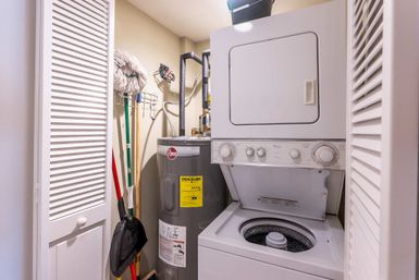 Small laundry utility closet with white stacked washer-dryer (top-loading washer open), gray water heater, mop, broom and dustpan stored behind louvered doors