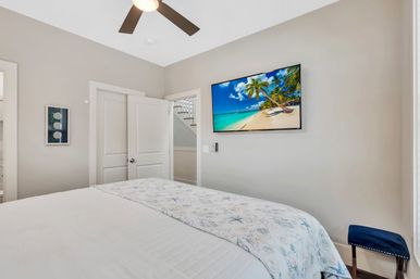 Bright beach-house bedroom with white bed and starfish-print coverlet, ceiling fan, wall-mounted TV showing a tropical beach, open door to stairs, neutral walls and a small blue stool.
