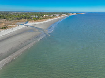 Aerial view of a sweeping curving sandy beach with a tidal inlet flowing into calm turquoise ocean, coastal dunes and trees under a clear blue sky.