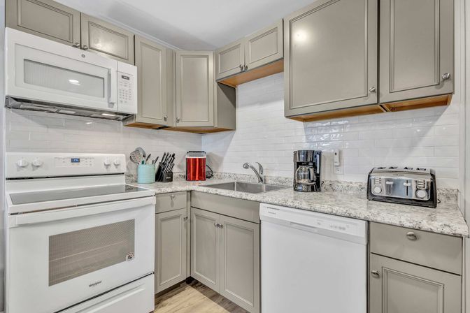 Bright modern kitchen with gray cabinets, white range and microwave, speckled granite countertops, white subway tile backsplash, stainless sink, coffee maker, toaster, and knife block.