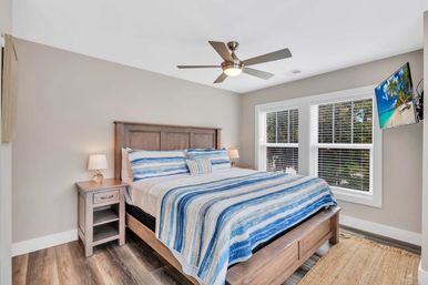 Sunlit coastal-style bedroom with wooden king bed, blue-and-white striped quilt and pillows, matching nightstand with lamp, ceiling fan, large double windows with white blinds, wall-mounted TV and hardwood floor.