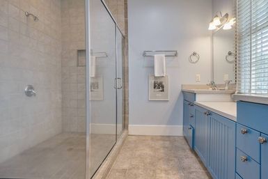Bright modern bathroom with glass-enclosed tiled walk-in shower, cheerful blue vanity with sink and countertop, neutral tile floor, wall-mounted towel rack, mirror and window blinds letting in natural light.