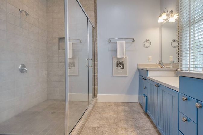 Bright modern bathroom with glass-enclosed tiled walk-in shower, cheerful blue vanity with sink and countertop, neutral tile floor, wall-mounted towel rack, mirror and window blinds letting in natural light.