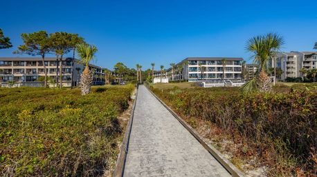 Sunny beachfront boardwalk flanked by dune vegetation and palm trees, leading to low-rise coastal condominiums under a clear blue sky.