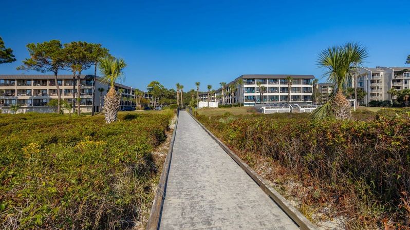 Sunny beachfront boardwalk flanked by dune vegetation and palm trees, leading to low-rise coastal condominiums under a clear blue sky.
